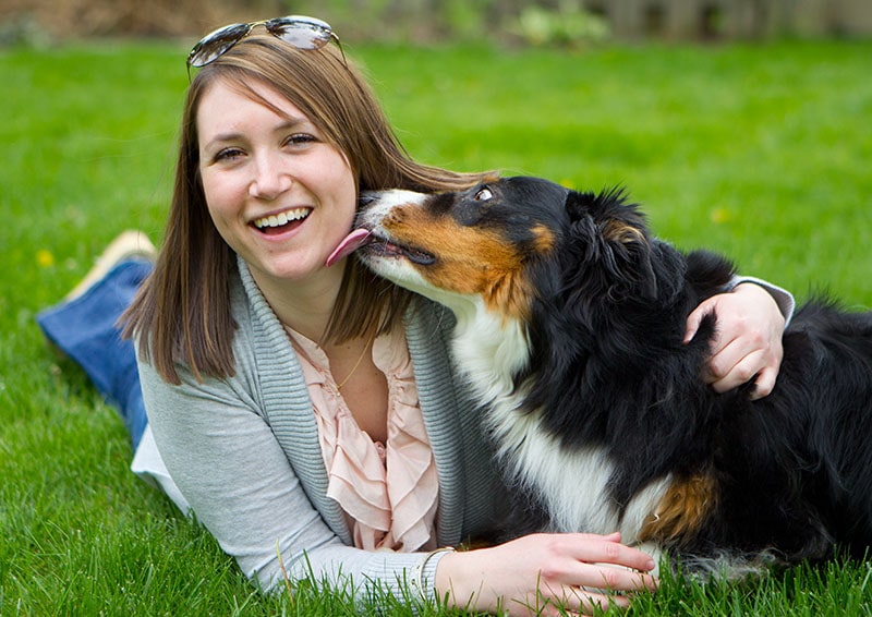 A happy and smiling women laying in the grass with her dog licking her face