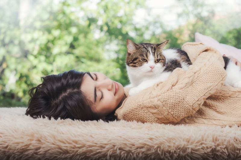 woman in brown sweater lay down on fluffy brown carpet with cat over her chest