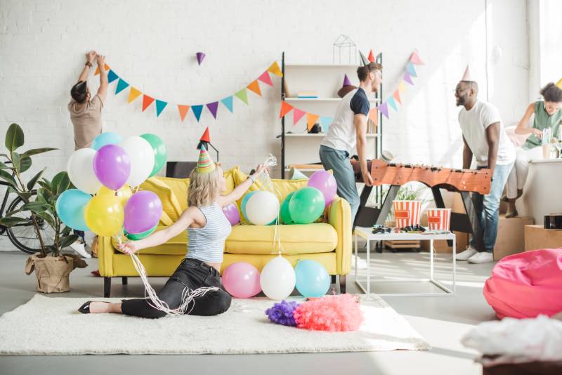 young woman sitting on floor with colorful balloons and her friends decorating room behind