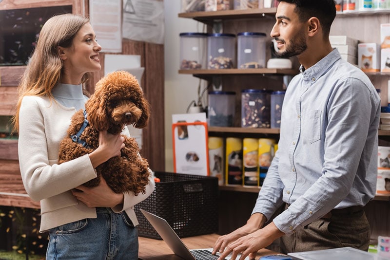 mujer sonriente sosteniendo un perro caniche y hablando con un hombre en una tienda de mascotas